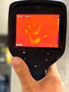 Eine Hand die eine Wärmebildkamera hält, ein verschwommener Hintergrund. Das Bild auf der Kamera zeigt schemenhaft eine gelbe Hand, die eine Stop-Geste zeigt und auf einem roten Hintergrund steht.